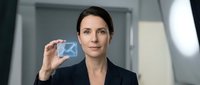 Woman with dark hair and fair complexion, appearing to be of European descent, wearing a black business blazer, holding up a translucent digital envelope icon in a modern office setting with soft lighting.