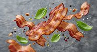 Crispy bacon strips flying through air with fresh basil leaves and peppercorns on dark stone background. Food photography for culinary advertising and menu design.