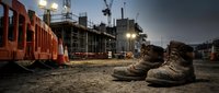 Pair of worn work boots on dusty ground at active construction site with cranes, scaffolding and industrial buildings in background, representing labor, safety and building industry.