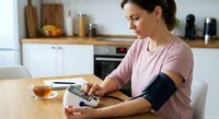 Young Caucasian woman measuring blood pressure at home using digital monitor in modern kitchen, healthcare self-monitoring and wellness routine.