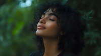Young African American woman with curly natural hair and serene expression against green blurred nature background, eyes closed in peaceful moment.