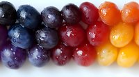 Fresh colorful grapes arranged in gradient rows from dark purple to orange yellow, with water droplets on glossy surface, creating rainbow fruit pattern.
