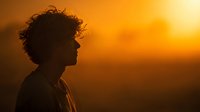 Silhouette of young person with curly hair against vibrant orange sunset sky, creating dramatic backlit profile in golden hour light.