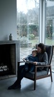 Mother and toddler son reading book together in armchair by rainy window near fireplace in cozy modern living room during stormy weather.