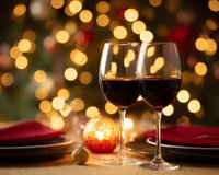 Two glasses of red wine on festive table setting with blurred golden bokeh lights and ornament, romantic holiday atmosphere for Christmas dinner celebration.