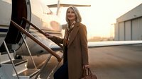 Middle-aged woman with blonde hair, Caucasian ethnicity, wearing brown coat and holding luggage, standing at private jet stairs during golden hour sunset. Business travel and luxury lifestyle concept.