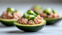 Fresh avocado halves filled with tuna salad and topped with diced cucumber, served on light background. Healthy keto diet appetizer for menu design.