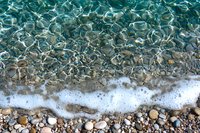 Crystal clear turquoise water meets pebbled beach shore with white foam waves, creating natural abstract patterns in Mediterranean coastal landscape.