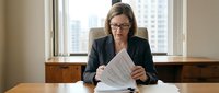 Professional businesswoman in glasses reviewing documents at office desk with city view background for corporate presentations.