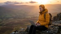 Woman in yellow jacket working on laptop while sitting on rocky mountain cliff at sunset, overlooking vast valley landscape for remote work and travel concepts.