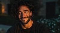 Handsome young Hispanic man with curly hair and beard smiling warmly in cozy evening lighting for lifestyle and portrait photography projects.