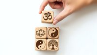 Hand placing wooden cube with Om symbol among spiritual blocks featuring lotus, yin yang and tree of life on white background for meditation design.