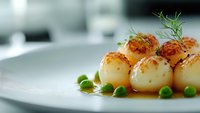 Pan-seared scallops garnished with fresh green peas and dill on white plate, served in caramelized sauce with selective focus for food photography.