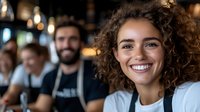 Young Mediterranean woman with curly hair and bright smile working as barista in modern coffee shop with coworkers in background, hospitality industry concept.