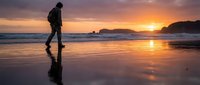 Silhouetted male traveler standing on wet beach sand during golden sunset with ocean waves and distant islands, perfect for travel and adventure themes.