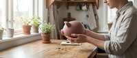 Young Caucasian man holding a pink piggy bank at a wooden kitchen table with coins, symbolizing savings, budgeting, and financial planning.