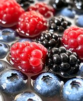 Fresh mixed berries with water drops in macro view showing juicy raspberries, blackberries and blueberries arranged in rows on light surface.