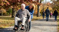 Caring adult daughter pushing elderly father in wheelchair through autumn park with colorful fall foliage. Family bonding and senior care concept.