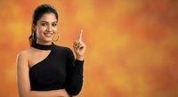 Young Indian woman in black one-shoulder top pointing upward with finger against warm orange gradient background for advertising and promotional content.