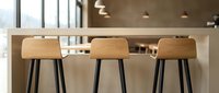 Young woman with light skin tone, wearing casual attire, sits at modern bar counter with three wooden stools featuring black metal legs. Minimalist kitchen interior with pendant lighting and neutral tones. Contemporary home design and hospitality setting.
