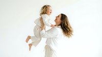Happy Caucasian mother in white robe lifting laughing toddler daughter up in air during playful bonding moment on clean white background.