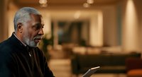 Senior African American man with gray hair wearing dark blazer, holding tablet device, sitting in modern home interior with warm lighting and blurred background, representing technology adoption, remote work, and contemporary lifestyle.