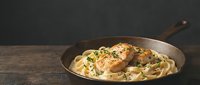 Grilled chicken breast with creamy mushroom pasta in cast iron skillet on rustic wooden table, gourmet dinner meal ready to serve.
