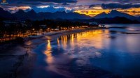 Coastal beach at twilight with golden streetlights reflecting on wet sand and calm ocean waters, dramatic mountain silhouettes against colorful sunset sky, perfect for travel and nature themes.