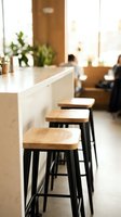 Modern cafe counter with wooden bar stools and white countertop, featuring blurred background with customers and plants, suitable for hospitality and restaurant design projects.