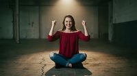 Young woman celebrating success sitting cross-legged on floor in urban loft space with raised fists showing joy and achievement in casual red top.