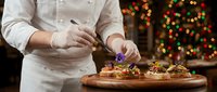 Professional chef in white uniform garnishing gourmet holiday appetizers on wooden board with festive Christmas lights background for restaurant menu.