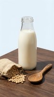 Fresh soy milk in glass bottle with soybeans spilling from burlap sack and wooden spoon on dark wood cutting board against white background.