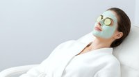 Young Caucasian woman relaxing with green clay facial mask and cucumber slices on eyes, wearing white robe at spa salon.