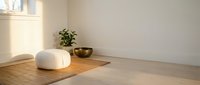 Minimalist meditation room with white cushion and wooden bowl on floor, natural light streaming through window creating peaceful zen atmosphere for mindfulness practice.