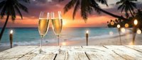Two champagne flutes on a rustic wooden table at a tropical beach sunset, with palm trees, tiki torches, and golden ocean waves in the background.