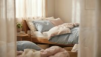 Cozy bedroom interior with unmade bed featuring soft pastel bedding, wooden nightstand, and natural sunlight streaming through sheer curtains, creating a warm and inviting sleeping space.