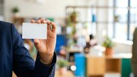 Male professional in dark business suit holding blank white business card at modern office workplace with blurred colleagues and plants in background for corporate branding and networking purposes.