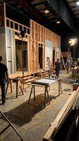 Film production set with wooden house facade under studio lights with crew members working on movie or television show.