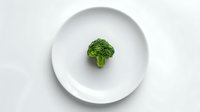 Fresh green broccoli floret centered on round white plate against minimalist white background, highlighting healthy eating and portion control.
