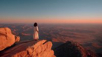 Woman in white dress standing on red rock cliff overlooking desert canyon landscape at golden hour sunset with dramatic mountain vista for meditation concepts.
