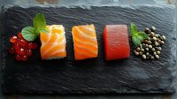 Fresh sushi ingredients arranged in row on black slate: red currants, salmon, tuna pieces, mint leaves and capers. Gourmet seafood composition for menu design.