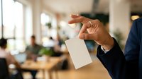 Male professional in business attire holding blank white card in modern office environment with colleagues working at desks in background, suitable for corporate communication and business messaging.