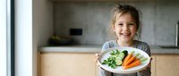 Happy young girl holding plate of fresh vegetables in modern kitchen, promoting healthy eating habits and nutrition education for children.