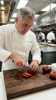 Mature male chef with gray hair and Mediterranean ethnicity, wearing white chef's coat, slicing fresh tomatoes on wooden cutting board in professional kitchen with staff working in background.
