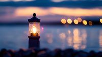 Glowing lighthouse lantern at dusk with blurred bokeh lights reflecting on calm water surface. Atmospheric maritime scene with warm illumination at sunset.