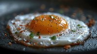 Sunny side up egg with vibrant orange yolk on dark surface, seasoned with fresh herbs, pepper and spices, captured in moody lighting with selective focus.