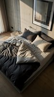 Modern bedroom with unmade bed featuring black and beige linen bedding, decorative pillows, wooden floor, and natural sunlight streaming through window for interior design projects.
