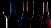 Wine bottles silhouetted against dark background with dramatic side lighting highlighting their elegant shapes and varied colors.