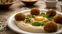 Fresh homemade falafel balls served on creamy hummus with herbs and olive oil drizzle on white plate, traditional Middle Eastern cuisine ready to eat.