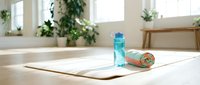 Yoga mat with water bottle and towel on wooden floor in bright studio space with plants and natural light.